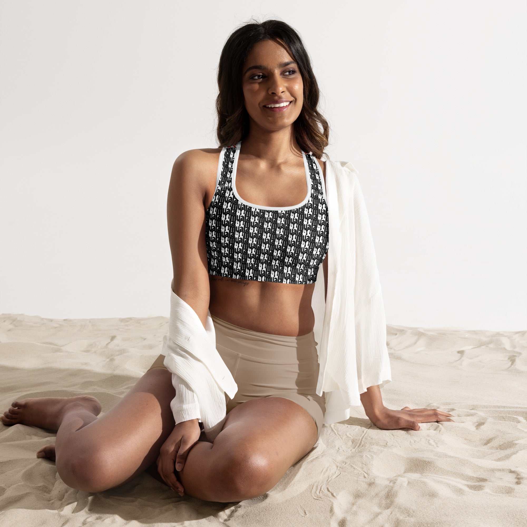 Woman wearing black and white patterned workout fuel sports bra sitting on sand with white cover-up draped over shoulders